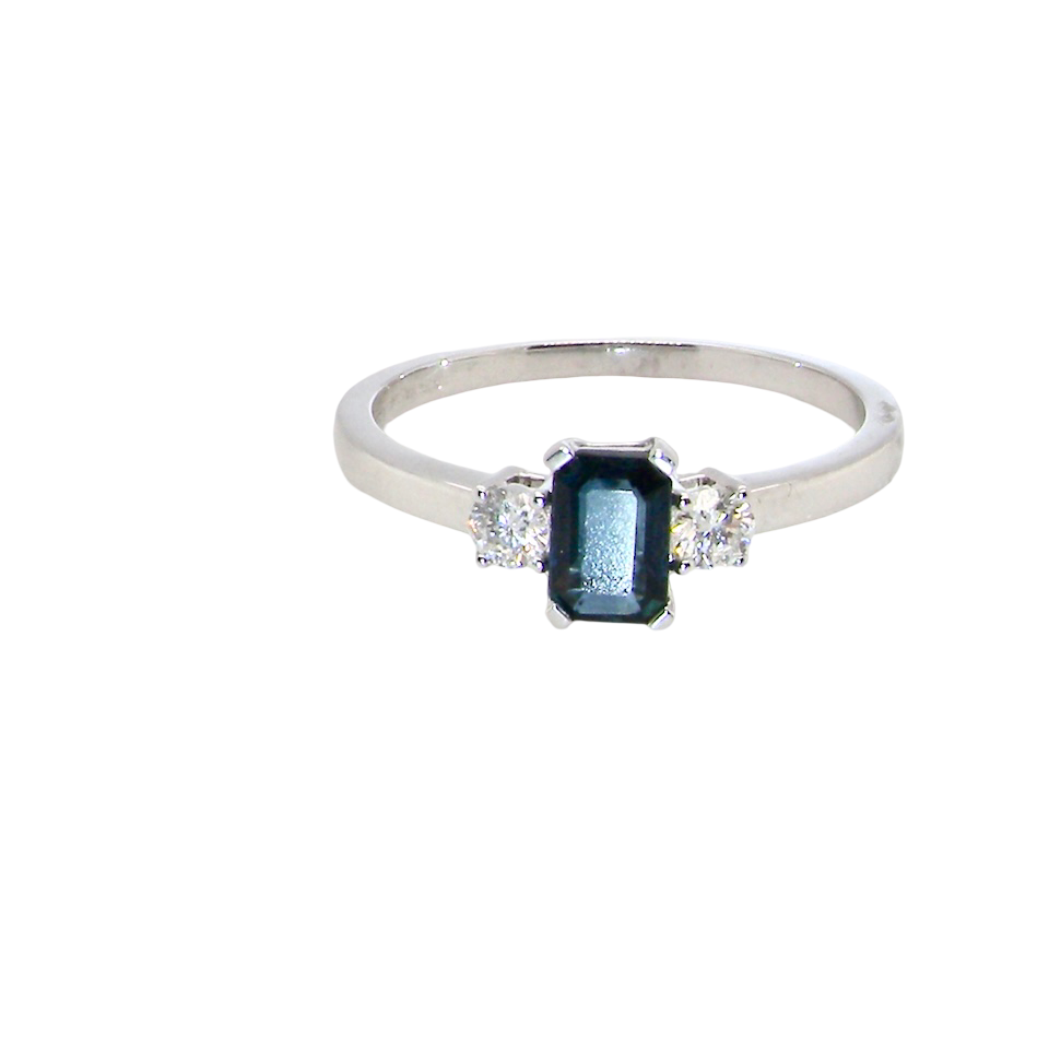 An elegant three-stone ring crafted in 18 carat white gold, featuring a striking rectangular dark blue sapphire at the centre, measuring 6 × 4 mm and weighing 0.50 carats.