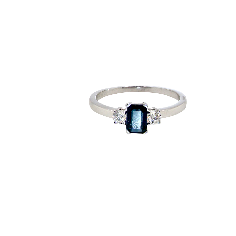 An elegant three-stone ring crafted in 18 carat white gold, featuring a striking rectangular dark blue sapphire at the centre, measuring 6 × 4 mm and weighing approximately 0.50 carats.