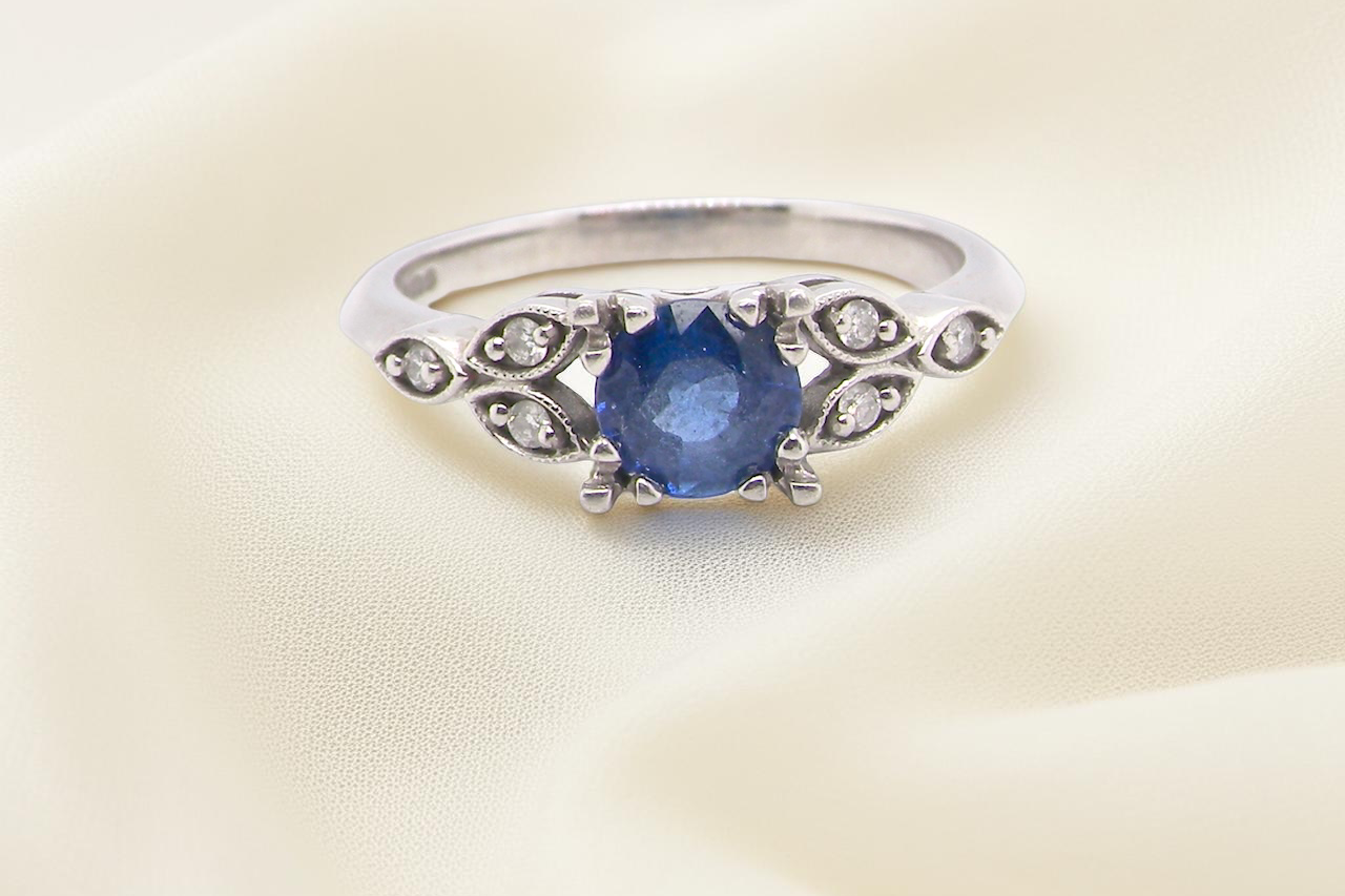 An elegant vintage-style platinum ring showcasing a beautifully proportioned round sapphire with a soft mid to light cornflower blue hue. The sapphire measures 5.6mm in diameter