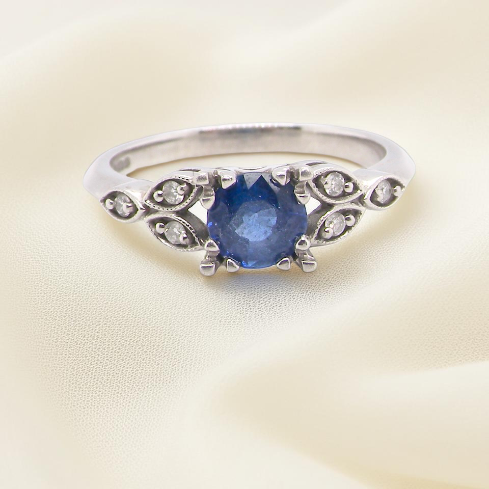 An elegant vintage-style platinum ring showcasing a beautifully proportioned round sapphire with a soft mid to light cornflower blue hue. The sapphire measures 5.6mm in diameter