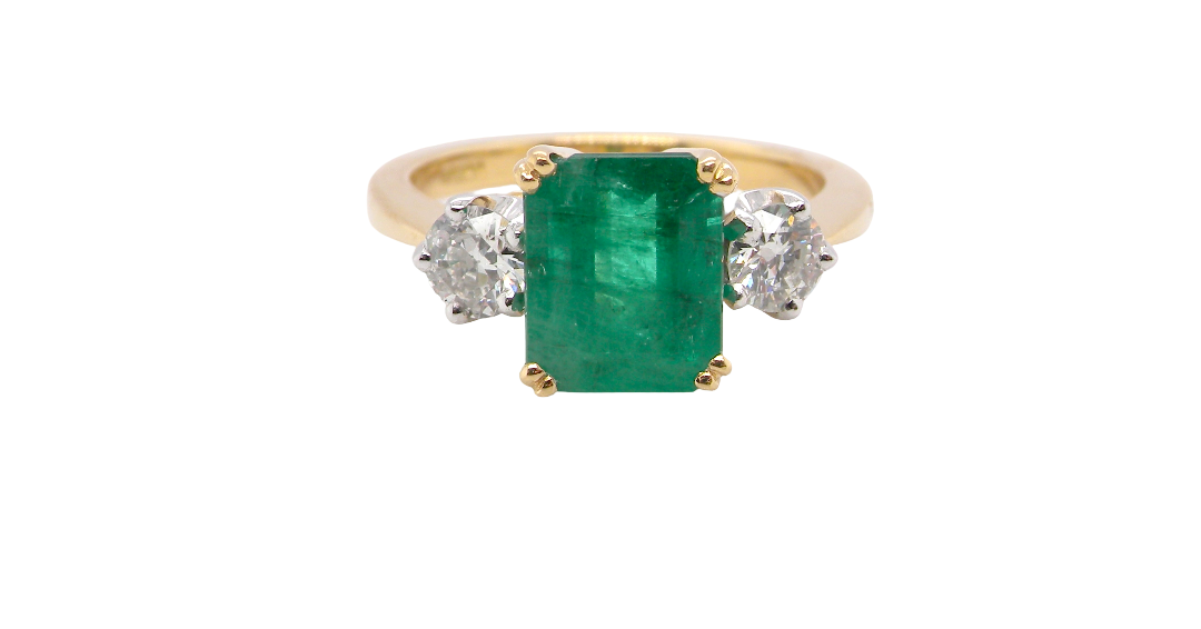 An exceptional three-stone ring featuring a vibrant step-cut emerald to the centre, measuring 8.6 x 7.0mm and weighing approximately 2.10 carats. The emerald displays a beautiful grass-green colour