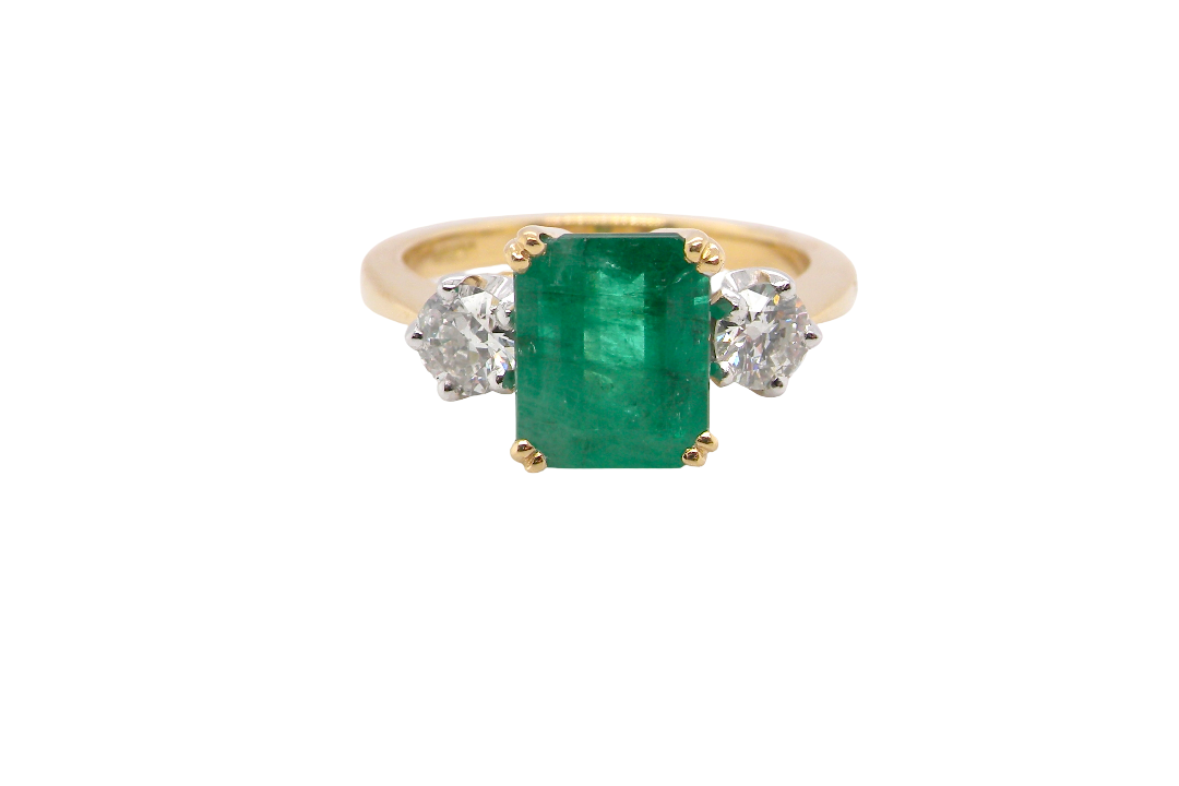 An exceptional three-stone ring featuring a vibrant step-cut emerald to the centre, measuring 8.6 x 7.0mm and weighing approximately 2.10 carats. The emerald displays a beautiful grass-green colour