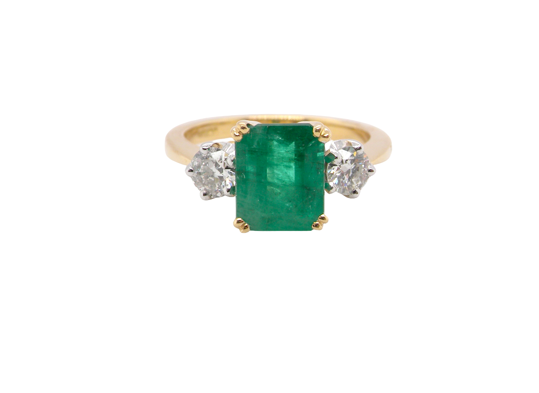 An exceptional three-stone ring featuring a vibrant step-cut emerald to the centre, measuring 8.6 x 7.0mm and weighing approximately 2.10 carats. The emerald displays a beautiful grass-green colour