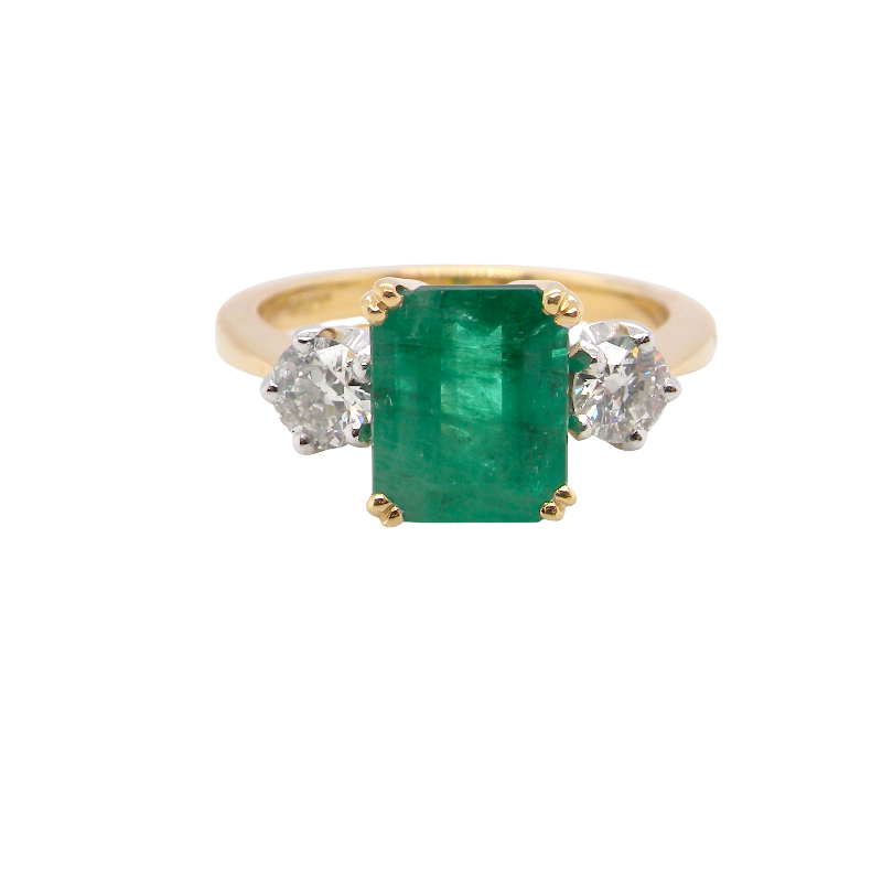 An exceptional three-stone ring featuring a vibrant step-cut emerald to the centre, measuring 8.6 x 7.0mm and weighing approximately 2.10 carats. The emerald displays a beautiful grass-green colour