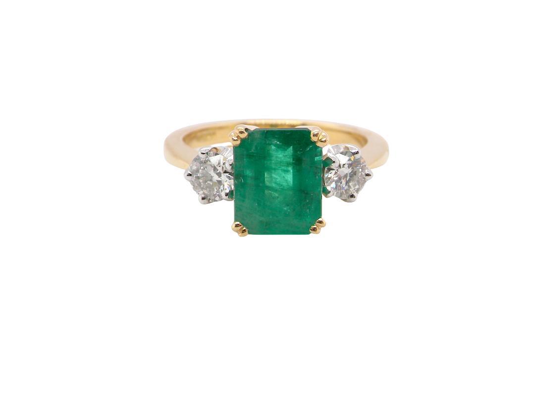 An exceptional three-stone ring featuring a vibrant step-cut emerald to the centre, measuring 8.6 x 7.0mm and weighing approximately 2.10 carats. The emerald displays a beautiful grass-green colour