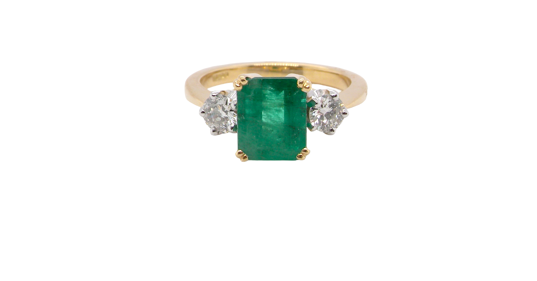 An exceptional three-stone ring featuring a vibrant step-cut emerald to the centre, measuring 8.6 x 7.0mm and weighing approximately 2.10 carats. The emerald displays a beautiful grass-green colour and is secured in a classic four-claw setting.