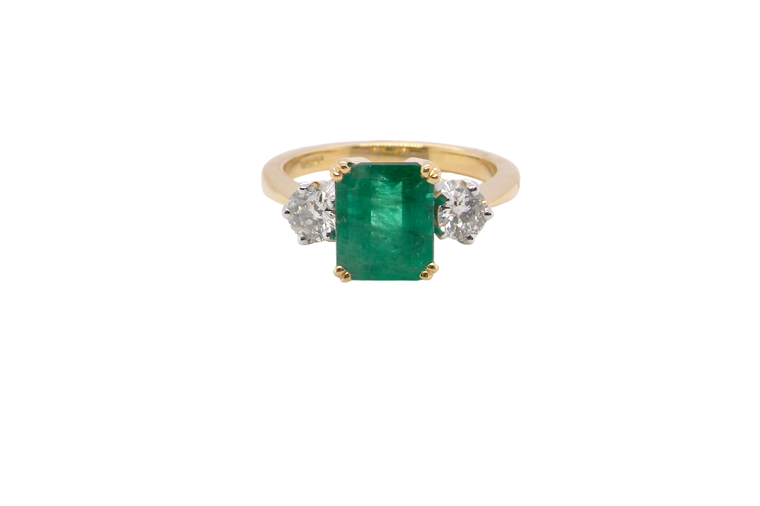 An exceptional three-stone ring featuring a vibrant step-cut emerald to the centre, measuring 8.6 x 7.0mm and weighing approximately 2.10 carats. The emerald displays a beautiful grass-green colour and is secured in a classic four-claw setting.
