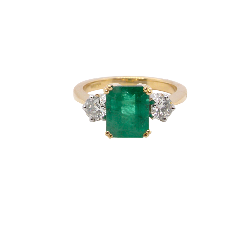 An exceptional three-stone ring featuring a vibrant step-cut emerald to the centre, measuring 8.6 x 7.0mm and weighing approximately 2.10 carats. The emerald displays a beautiful grass-green colour and is secured in a classic four-claw setting.