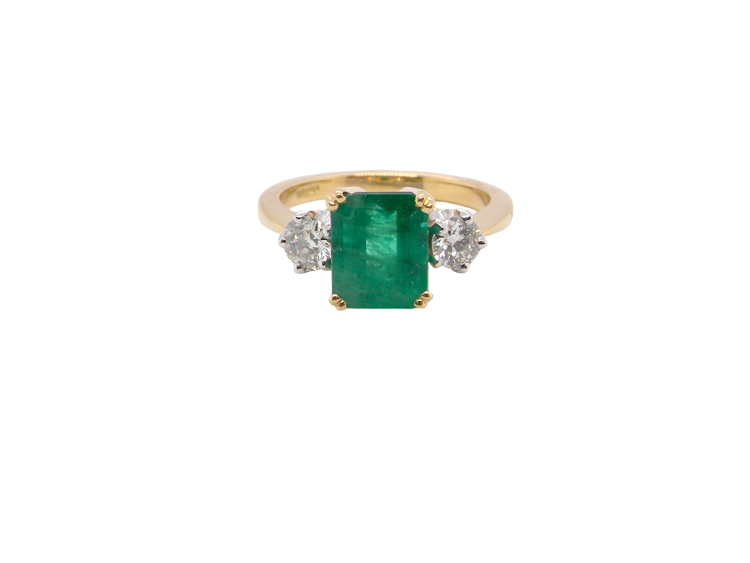 An exceptional three-stone ring featuring a vibrant step-cut emerald to the centre, measuring 8.6 x 7.0mm and weighing approximately 2.10 carats. The emerald displays a beautiful grass-green colour and is secured in a classic four-claw setting.