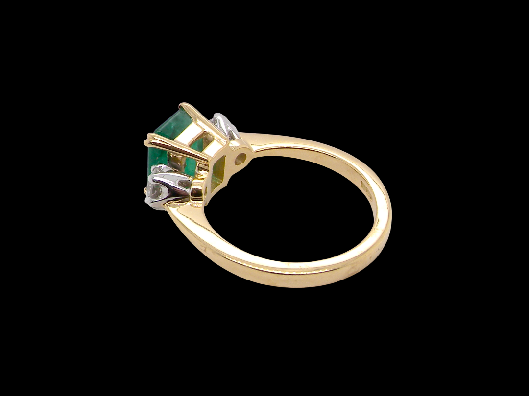 An exceptional three-stone ring featuring a vibrant step-cut emerald to the centre, measuring 8.6 x 7.0mm and weighing approximately 2.10 carats. The emerald displays a beautiful grass-green colour and is secured in a classic setting.
