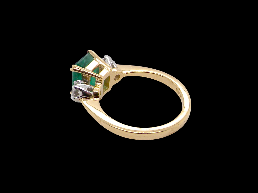 An exceptional three-stone ring featuring a vibrant step-cut emerald to the centre, measuring 8.6 x 7.0mm and weighing approximately 2.10 carats. The emerald displays a beautiful grass-green colour and is secured in a classic setting.