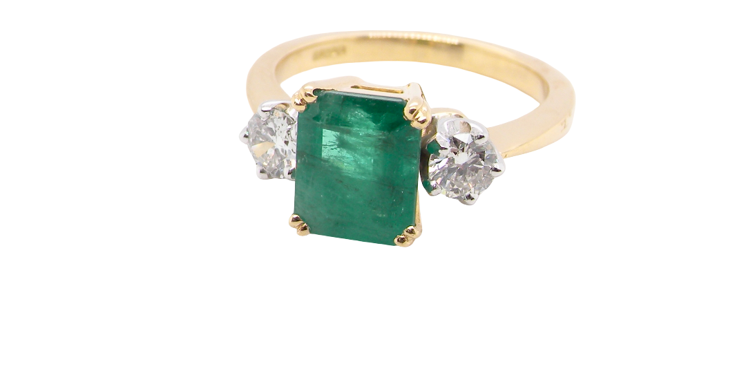 An exceptional three-stone ring featuring a vibrant step-cut emerald to the centre, measuring 8.6 x 7.0mm and weighing approximately 2.10 carats. The emerald displays a beautiful grass-green colour and is secured in a four-claw setting.