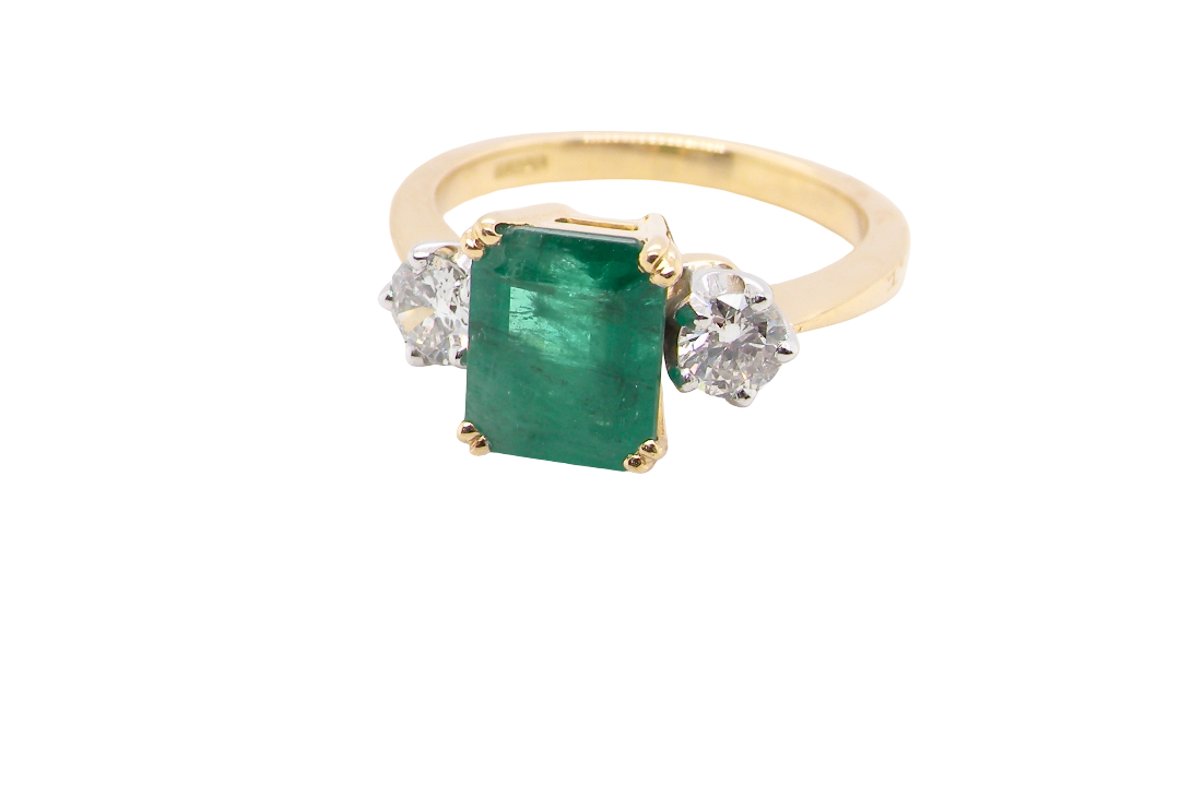 An exceptional three-stone ring featuring a vibrant step-cut emerald to the centre, measuring 8.6 x 7.0mm and weighing approximately 2.10 carats. The emerald displays a beautiful grass-green colour and is secured in a four-claw setting.