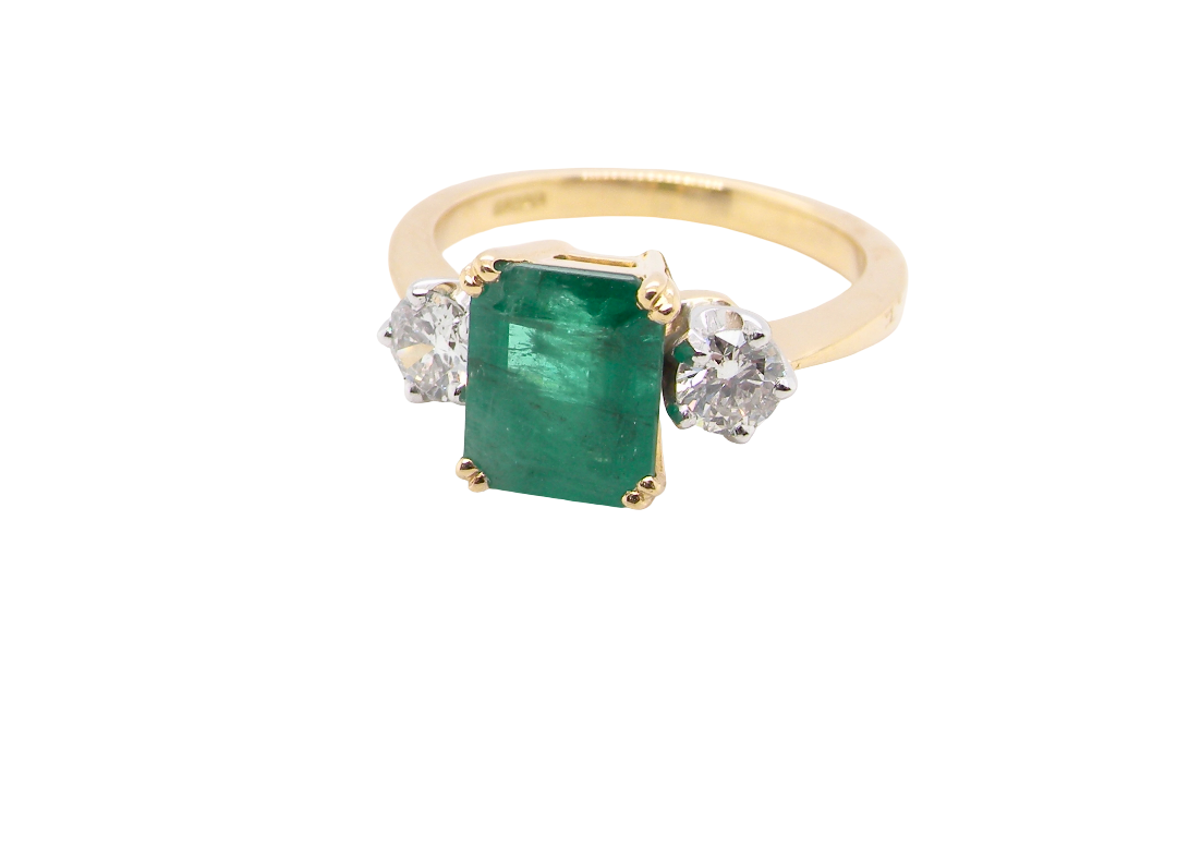 An exceptional three-stone ring featuring a vibrant step-cut emerald to the centre, measuring 8.6 x 7.0mm and weighing approximately 2.10 carats. The emerald displays a beautiful grass-green colour and is secured in a four-claw setting.
