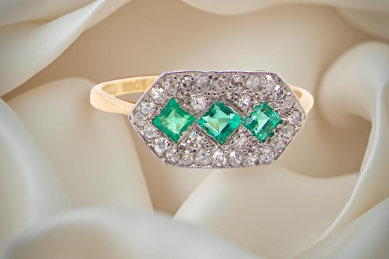 Edwardian era cluster ring, beautifully handcrafted with a trio of vibrant emeralds surrounded by a halo of antique old-cut diamonds.
