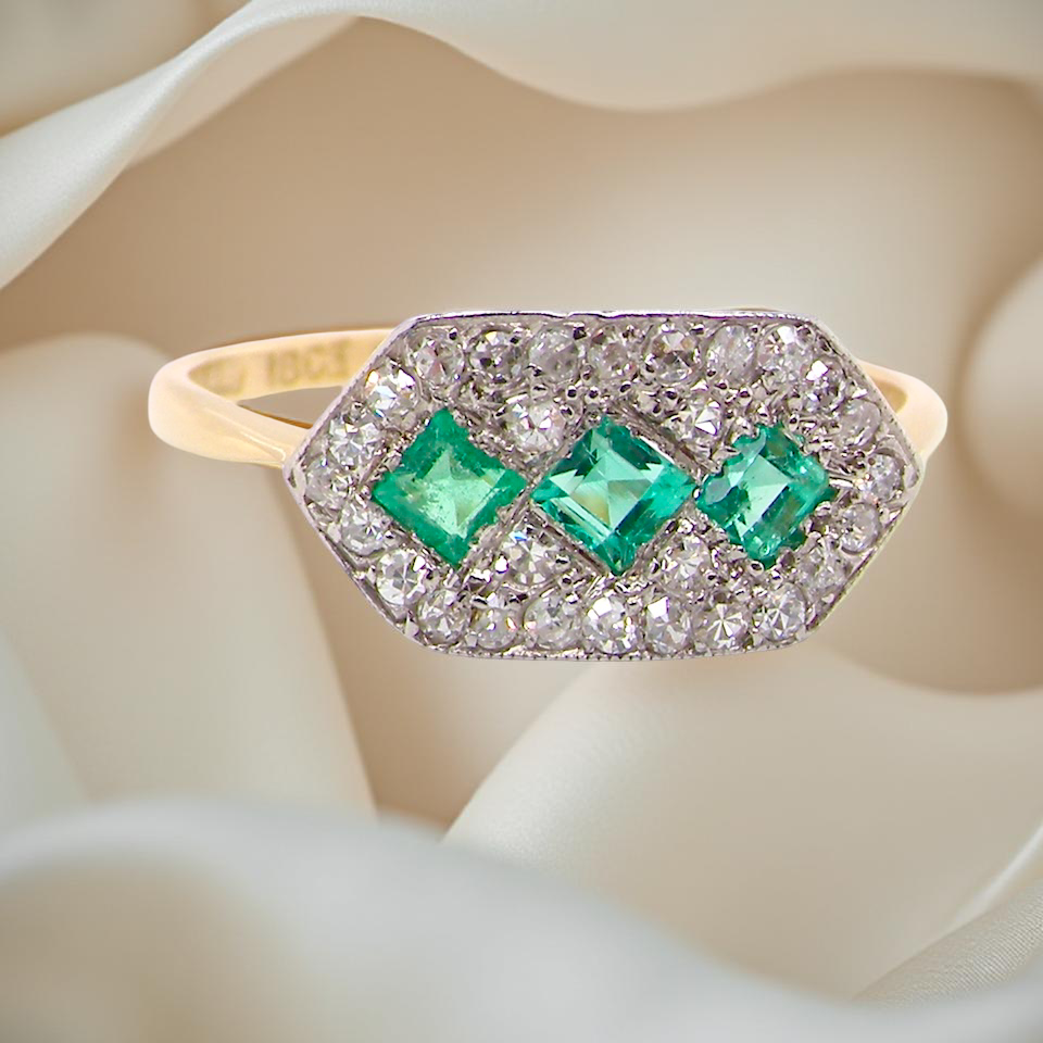 Edwardian era cluster ring, beautifully handcrafted with a trio of vibrant emeralds surrounded by a halo of antique old-cut diamonds.