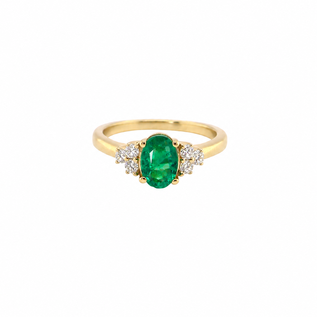 A yellow gold ring featuring an oval-cut green emerald center stone, flanked by small clusters of clear stones on either side, set against a plain white background.