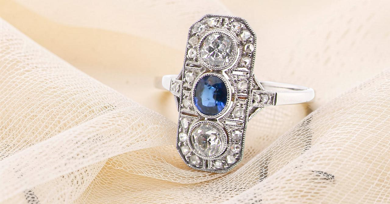 The plaque design has at its centre a mid to dark blue sapphire, 5x4mm (est 0.40 carats) mille grain set. Above and below to Old European Cut white diamonds.