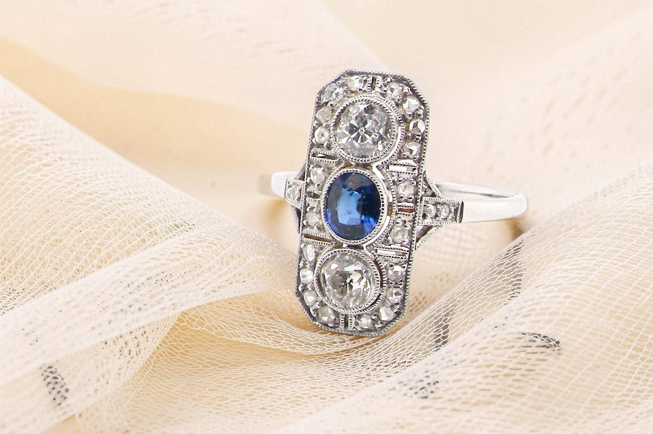 The plaque design has at its centre a mid to dark blue sapphire, 5x4mm (est 0.40 carats) mille grain set. Above and below to Old European Cut white diamonds.