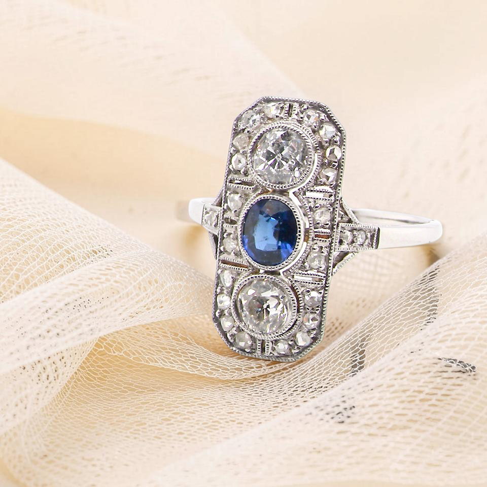 The plaque design has at its centre a mid to dark blue sapphire, 5x4mm (est 0.40 carats) mille grain set. Above and below to Old European Cut white diamonds.