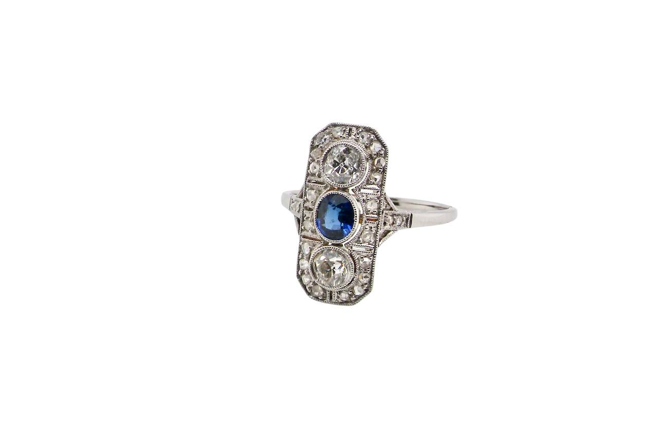 The plaque design has at its centre a mid to dark blue sapphire, 5x4mm (est 0.40 carats) mille grain set. Above and below to Old European Cut white diamonds. Each one estimated at 0.40 carats (4.8mm) Colour H-j, clarity Si.