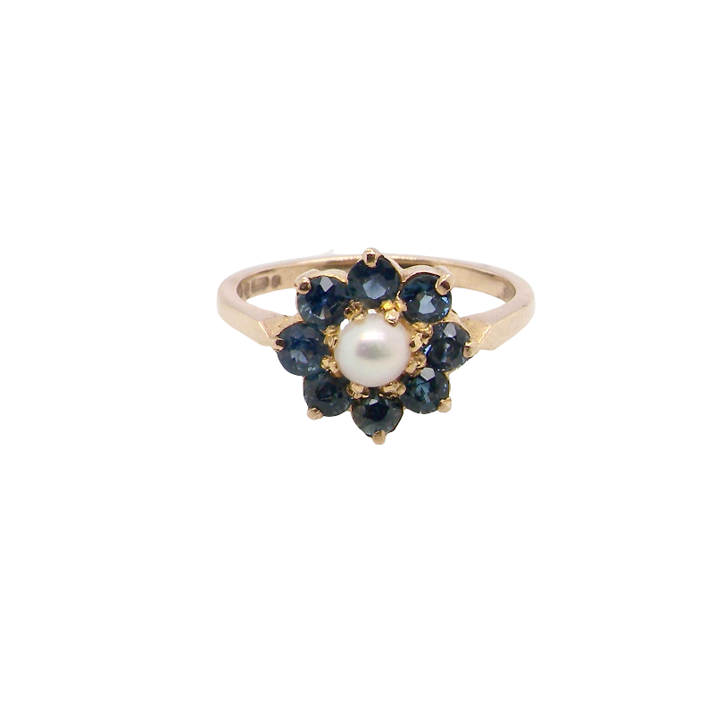 A vintage-style gold ring featuring a central white pearl surrounded by a halo of dark blue gemstones, set against a plain white background.