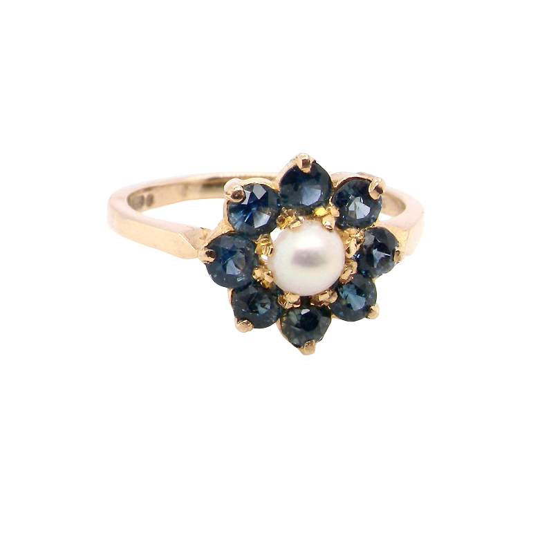 A yellow gold ring featuring a flower-shaped cluster with a central white pearl surrounded by dark blue round-cut gemstones, set against a plain white background.