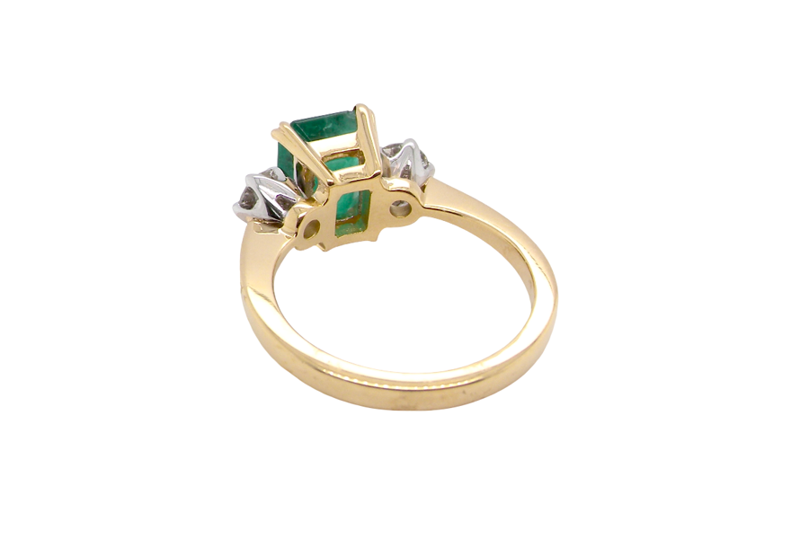 An exceptional three-stone ring featuring a vibrant step-cut emerald to the centre, measuring 8.6 x 7.0mm and weighing approximately 2.10 carats. The emerald displays a beautiful grass-green colour and is secured in a classic four-claw setting.