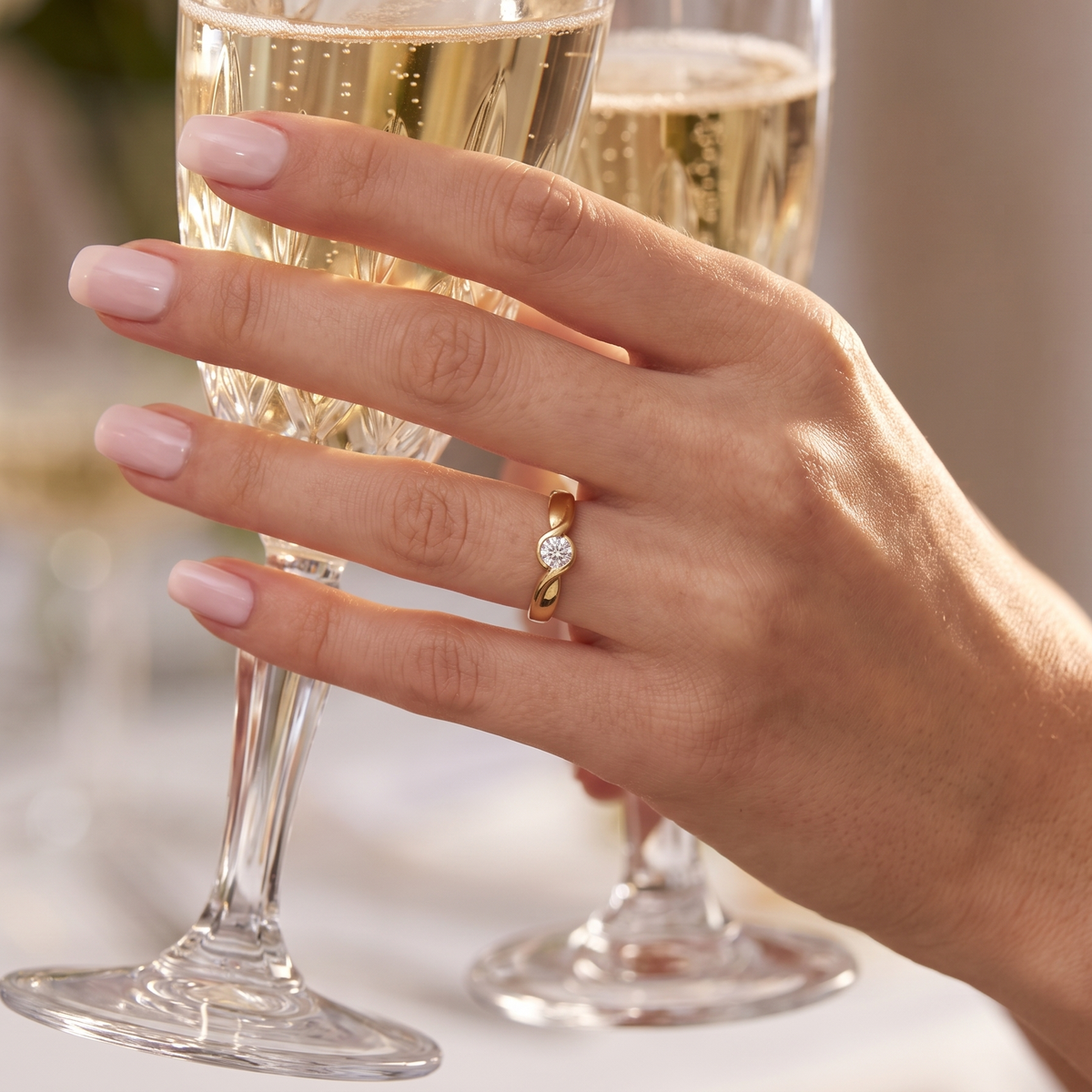 A close-up of a hand with pale pink manicured nails wearing a gold ring with a clear gemstone, holding a champagne flute in a softly lit indoor setting.
