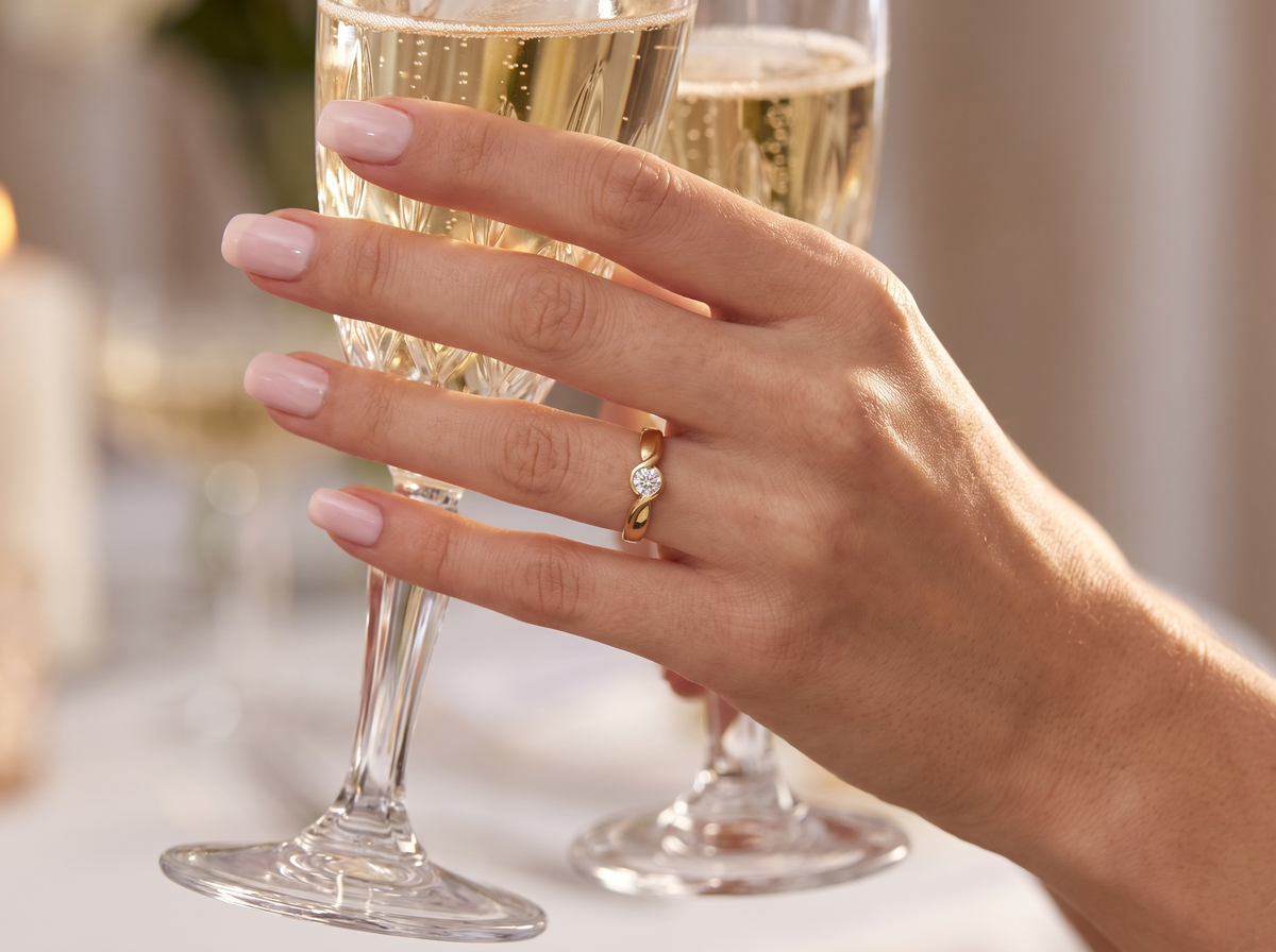 A close-up of a hand with pale pink manicured nails wearing a gold ring with a clear gemstone, holding a champagne flute in a softly lit indoor setting.