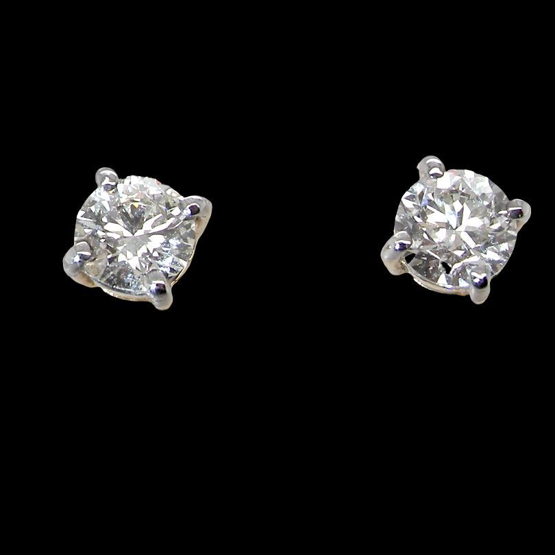 A pair of classic solitaire stud earrings featuring round brilliant-cut diamonds in four-prong silver settings, presented on a solid black background.