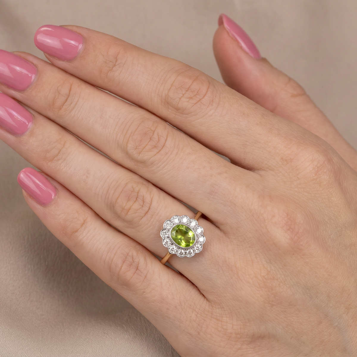A close-up of a hand with pink manicured nails wearing a gold ring featuring a green peridot stone surrounded by a halo of small clear stones, set against a soft beige fabric background.