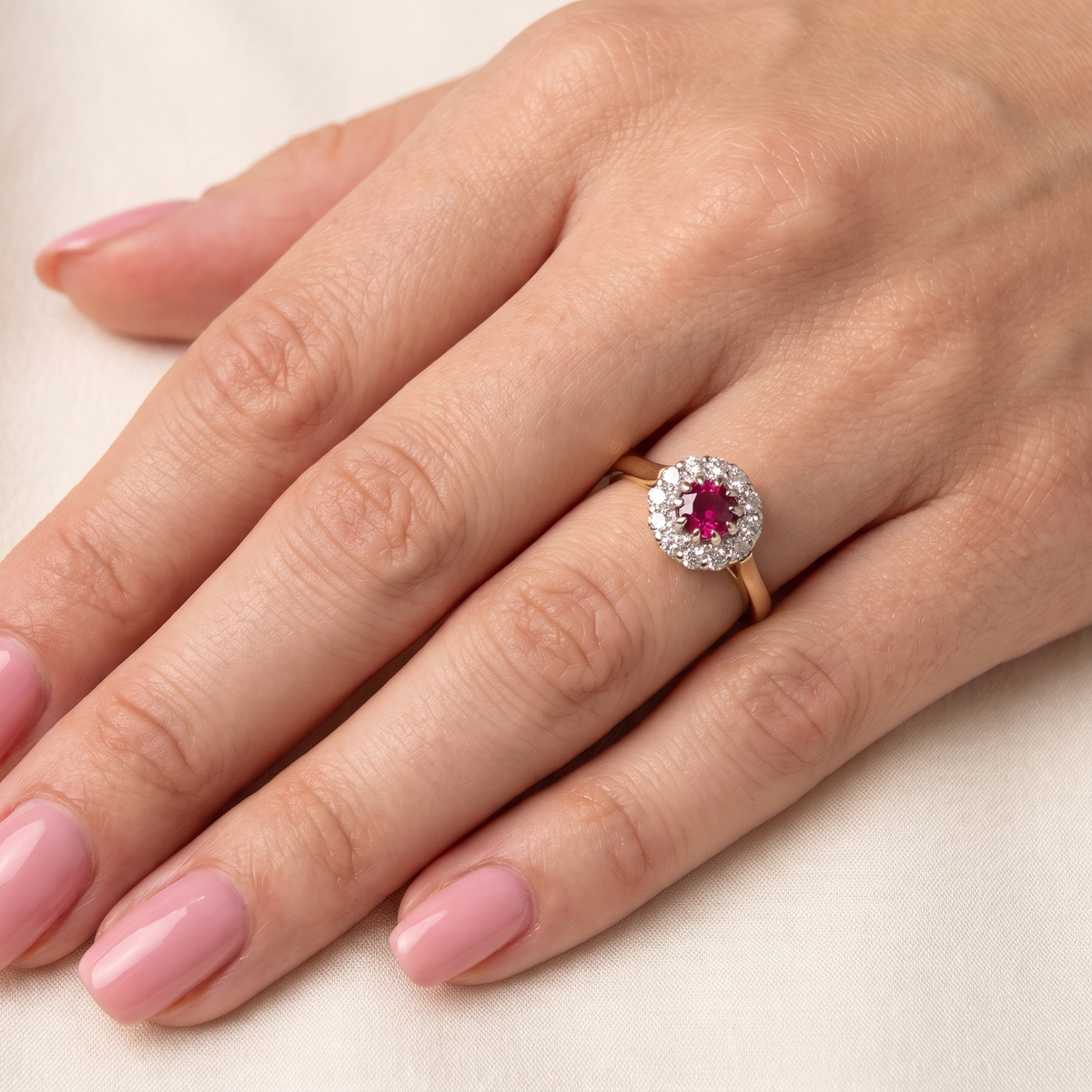 A close-up of a hand with pink manicured nails wearing a yellow gold ring featuring a round-cut pink gemstone surrounded by a halo of small clear stones, set against a soft cream fabric background.