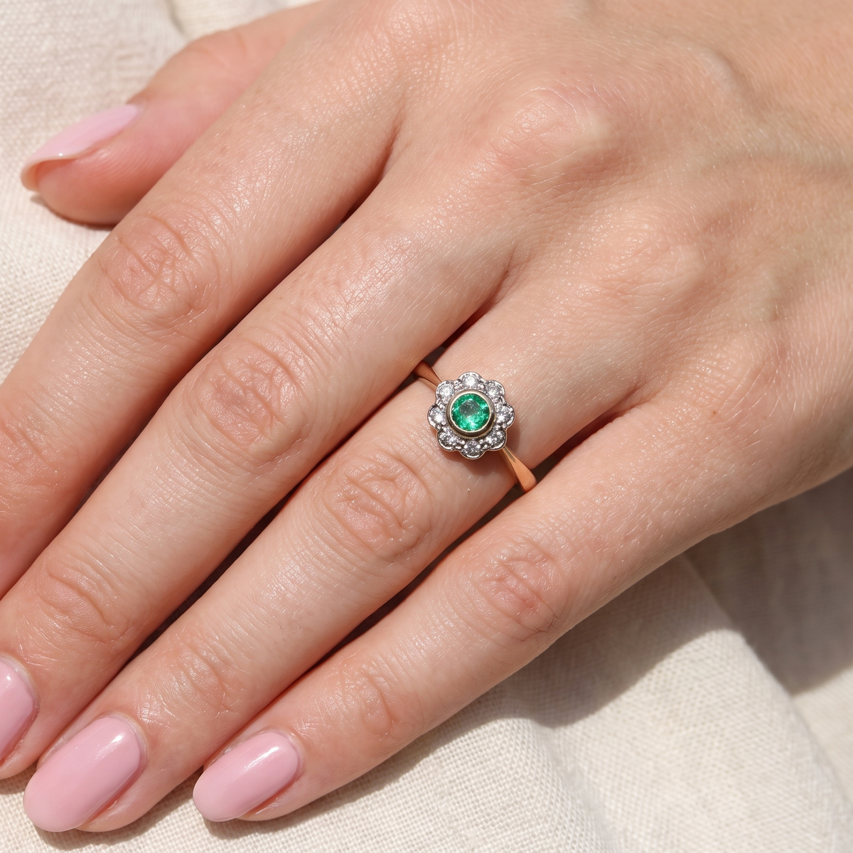 A close-up of a hand wearing a ring with a round green center stone surrounded by small clear stones, set on a rose gold band, resting on a light beige fabric background.