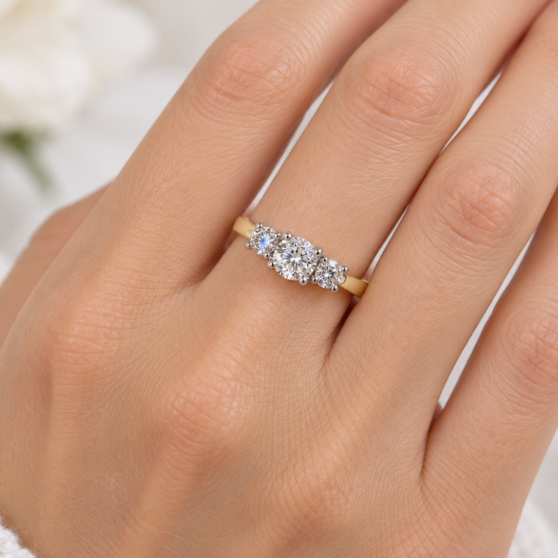 A close-up of a hand wearing a yellow gold three-stone ring with clear round-cut diamonds. The hand is shown against a soft, out-of-focus white background with a hint of green foliage.
