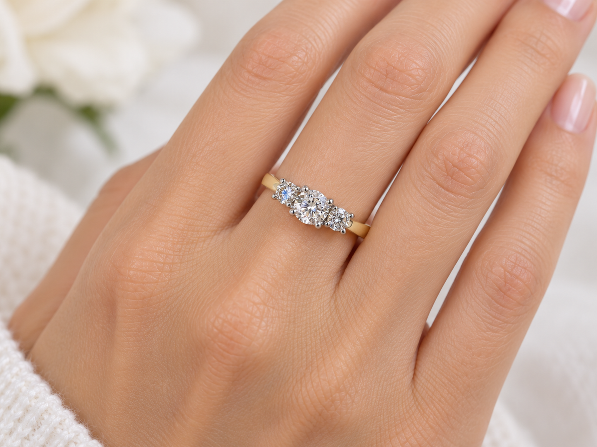 A close-up of a hand wearing a yellow gold three-stone ring with clear round-cut diamonds. The hand is shown against a soft, out-of-focus white background with a hint of green foliage.