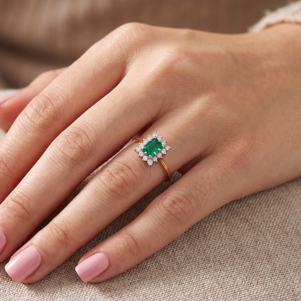 A close-up of a hand wearing a gold ring with a rectangular green emerald-cut center stone and small clear accent stones, resting on a beige textured fabric surface.