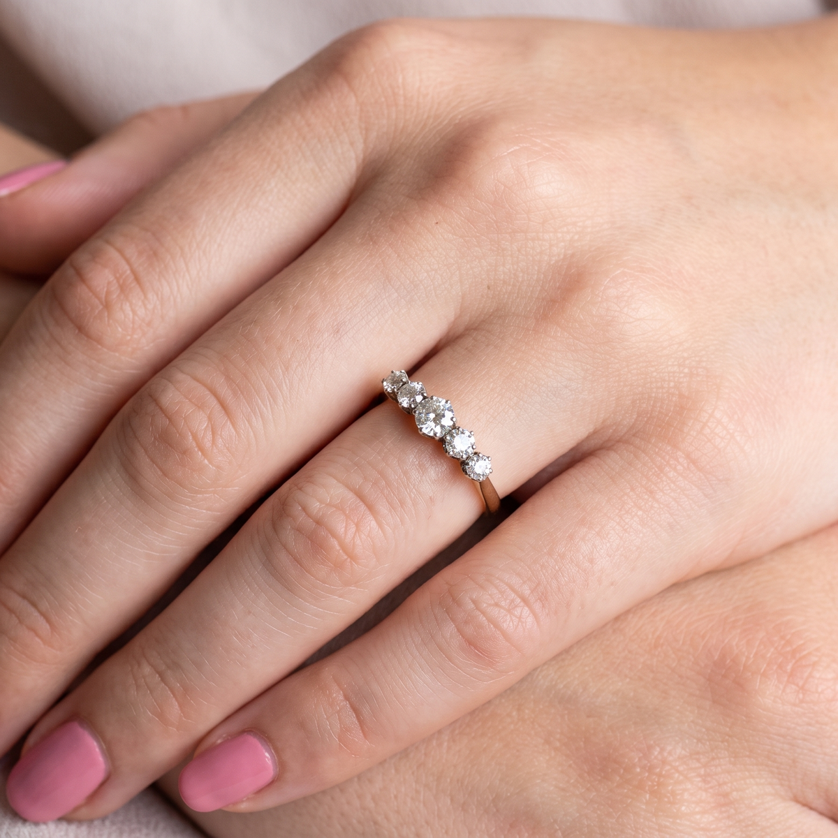 A close-up of a hand with pink manicured nails wearing a delicate gold ring set with small clear stones, resting on another hand against a soft neutral background.