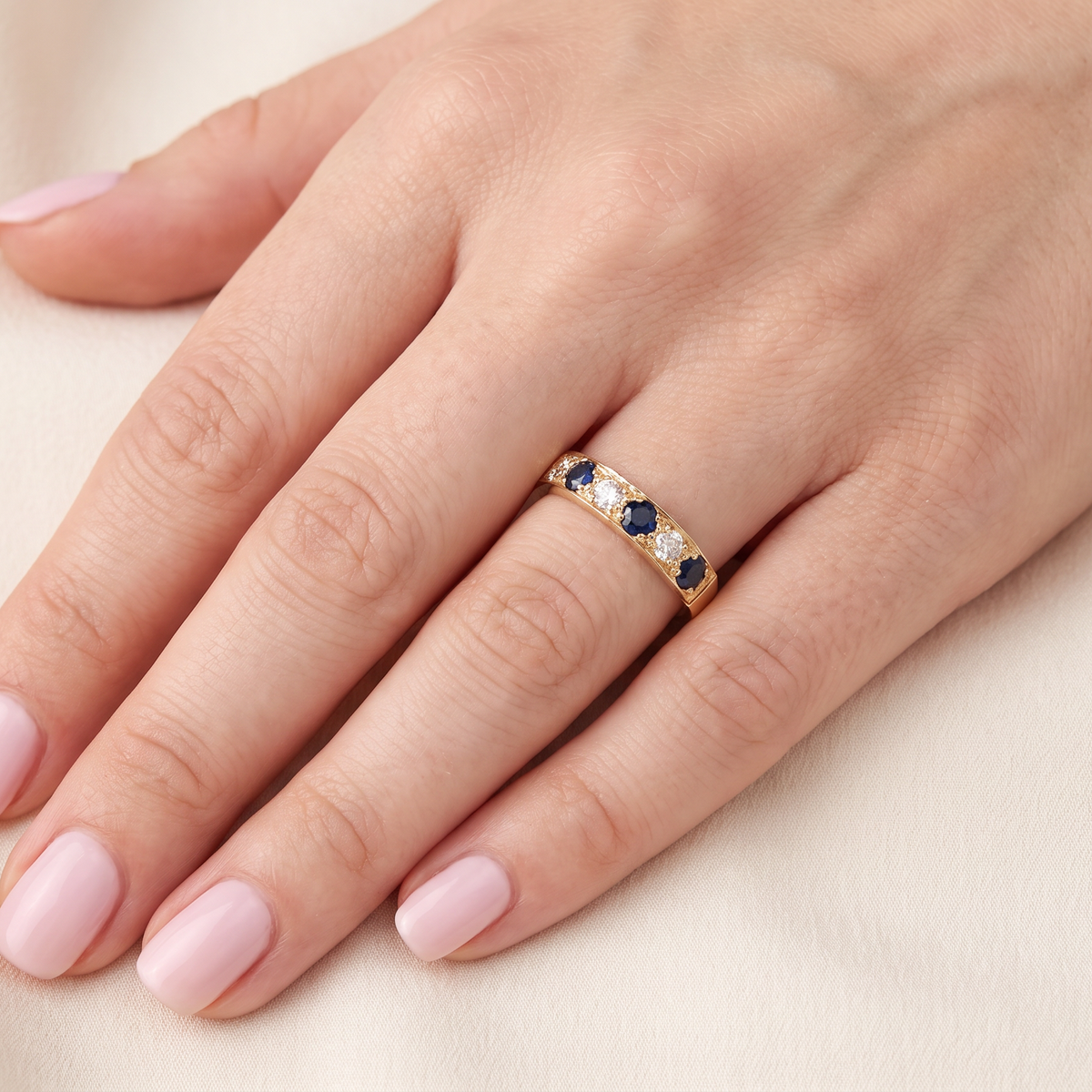 A close-up of a hand with light pink manicured nails wearing a gold band ring set with small blue and clear stones, resting on a soft cream fabric background.