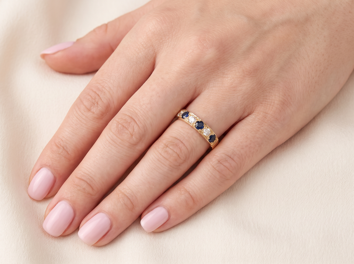 A close-up of a hand with light pink manicured nails wearing a gold band ring set with small blue and clear stones, resting on a soft cream fabric background.
