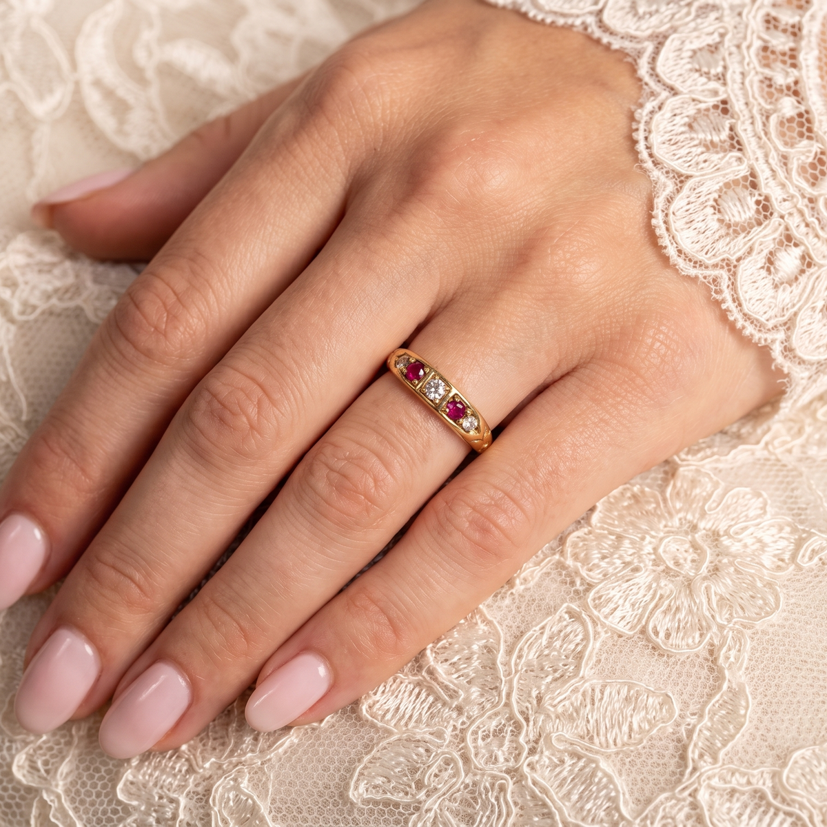 A close-up of a hand wearing a delicate gold band ring with small red and clear stones, resting on cream lace fabric.