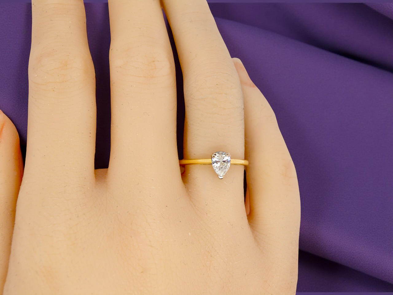Gold ring with a pear-shaped diamond on a hand against a purple fabric background