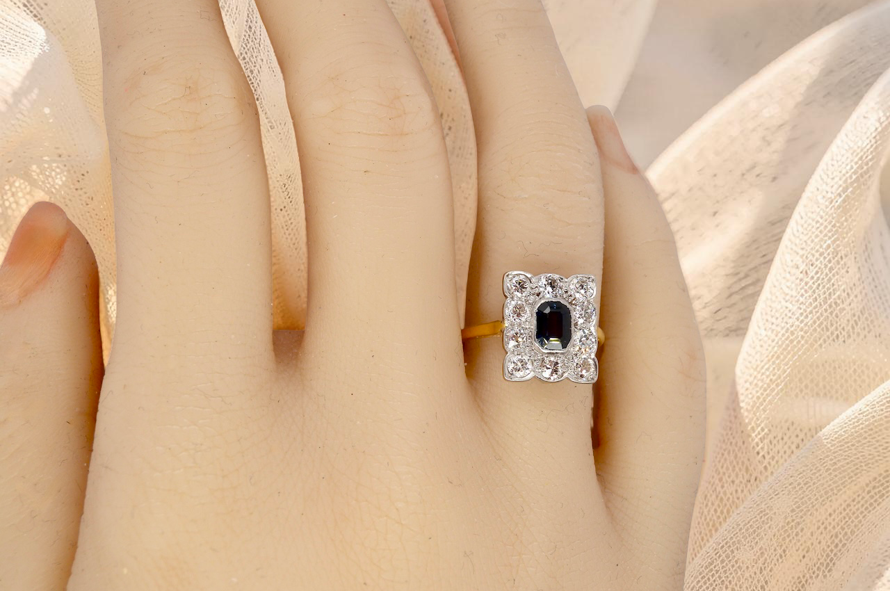 hand A rectangular cluster ring that features in its centre a cushion shaped dark blue sapphire, that measures 5x3.5mm (est 0.30 carats)