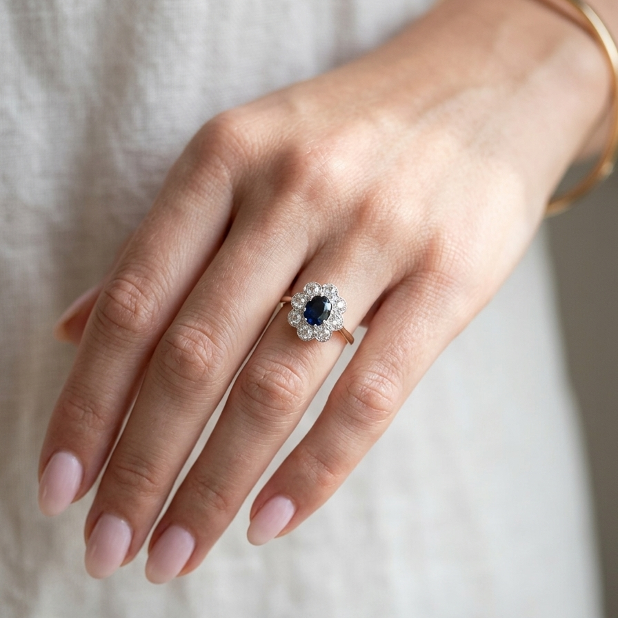hand A striking vintage sapphire and diamond cluster ring dating from the 1930s–1940s, beautifully crafted in platinum and 18 carat gold. At the centre sits a richly coloured oval-cut sapphire of deep inky blue-black hue, measuring approximately 7 x 5mm and estimated at 0.75 carats.