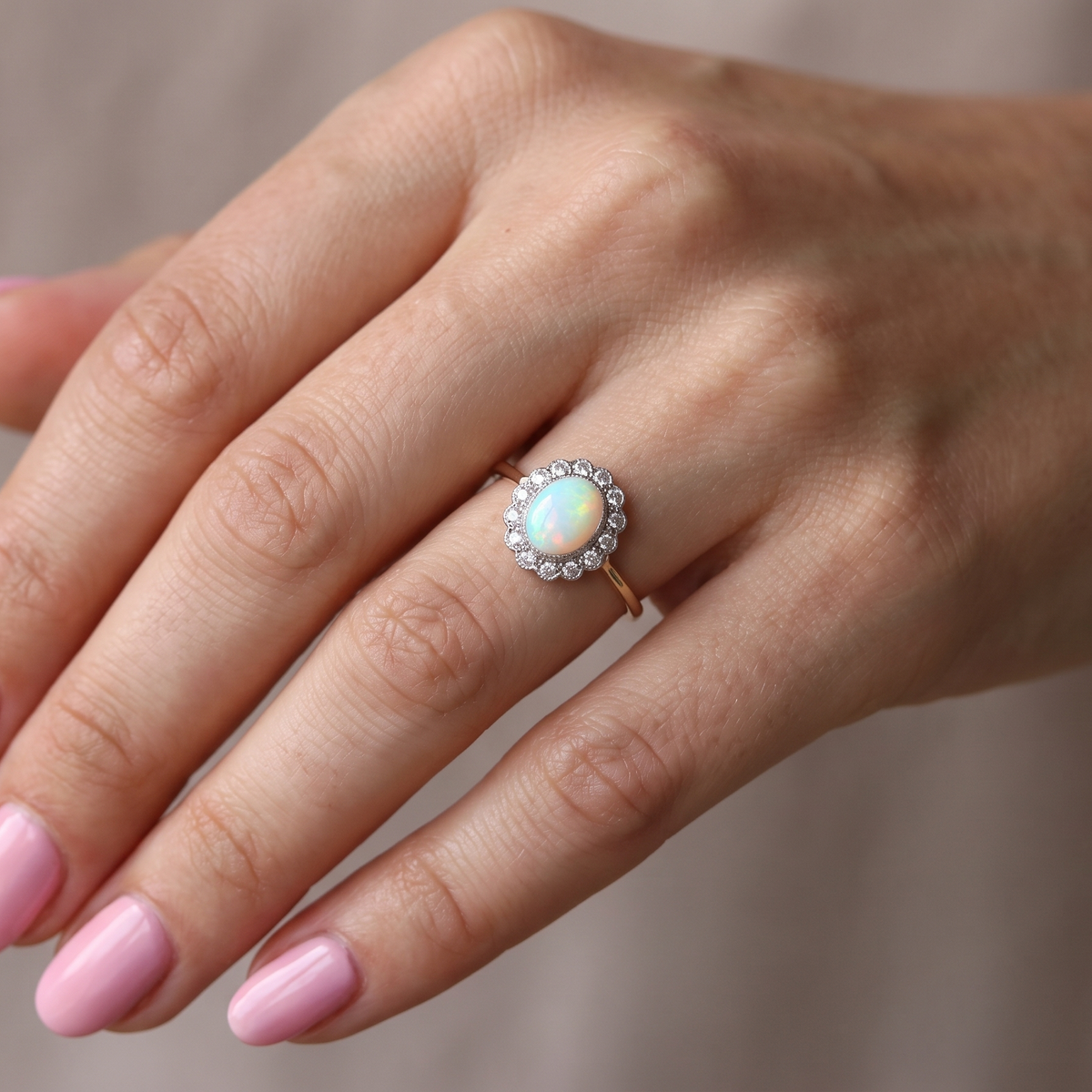 A close-up of a hand wearing a gold ring with a round iridescent opal center stone and a halo of small clear stones, set against a soft neutral background.