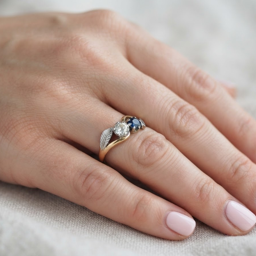 A close-up of a hand wearing a delicate ring with a gold leaf design, a clear center stone, and small dark blue accent stones, resting on a soft beige fabric background.