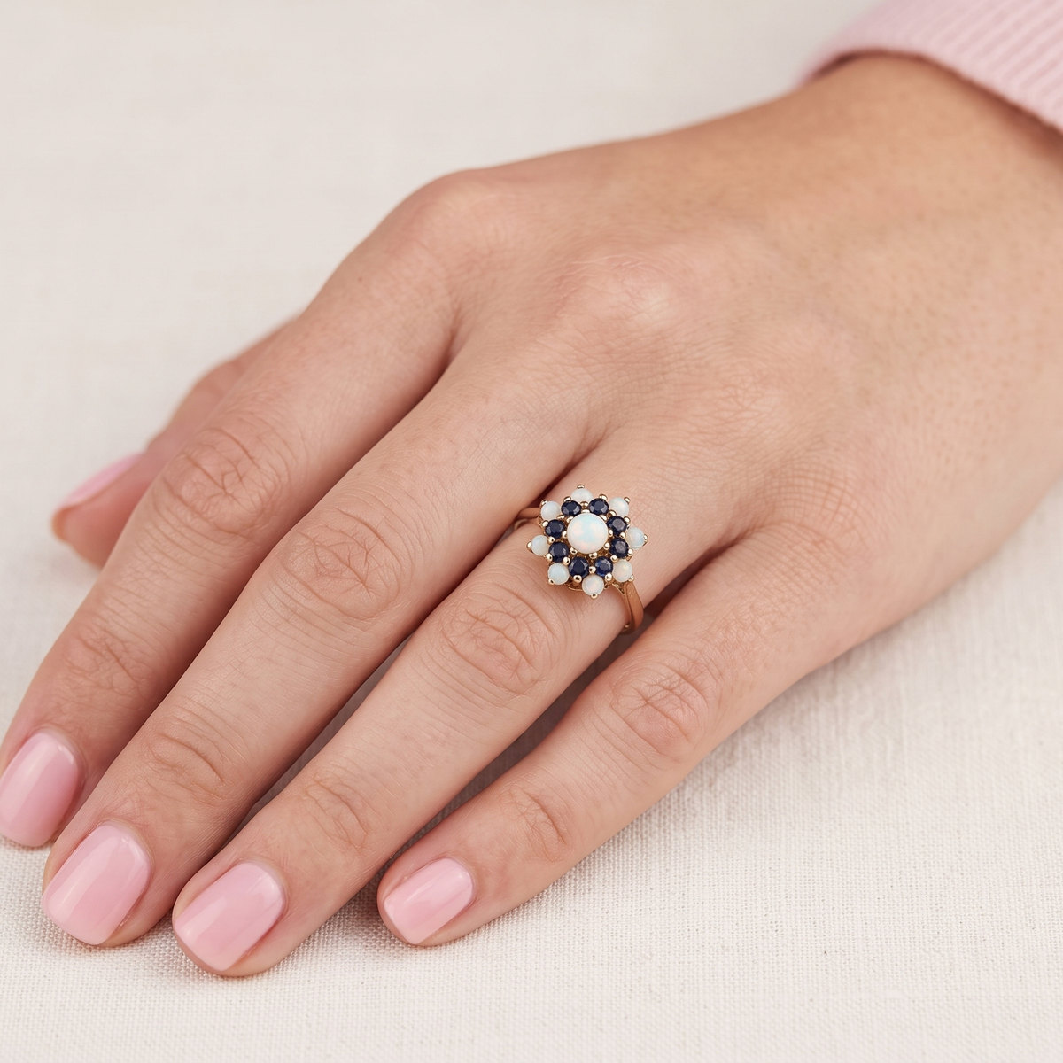A close-up of a hand wearing a small flower-shaped ring with a gold band, dark blue stones, and a central white stone, set against a light beige textured background.