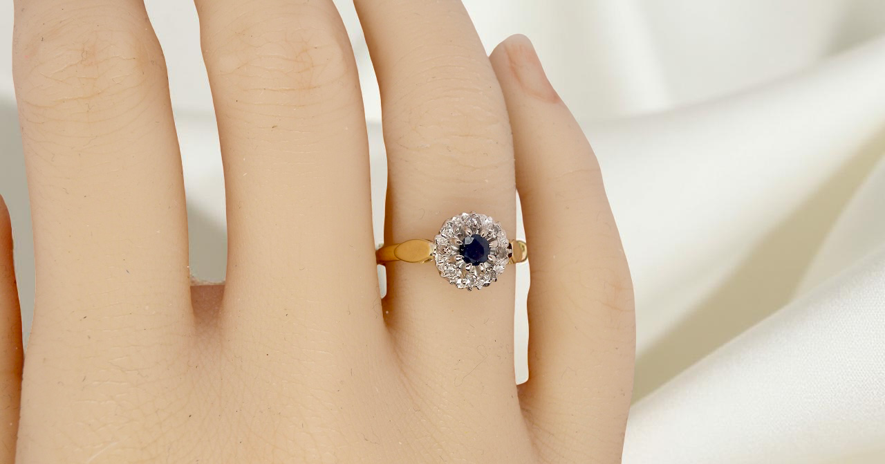 hand An 18 carat gold cluster ring, features to the centre a dark blue sapphire, 3.6mm diameter. Claw set.
