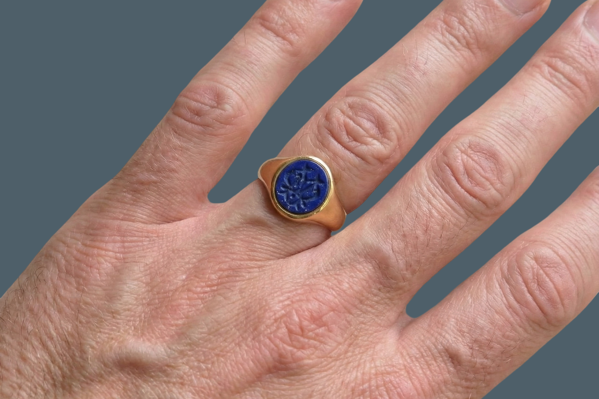 hand An 18 carat gold signet ring set with an oval lapis lazuli stone. This measures 12x9.5mm and has a carved intaglio seal engraved onto the stone. (These seem to be floral patterns)