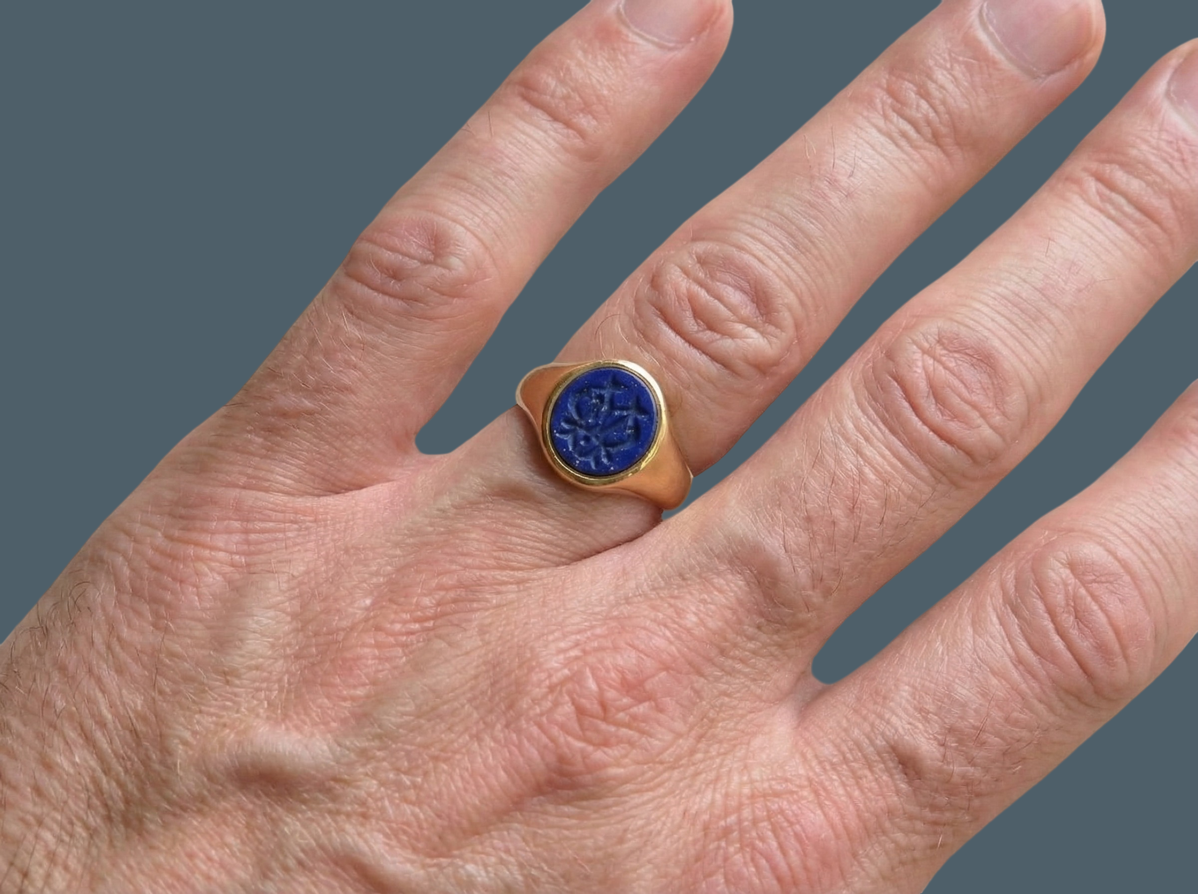 hand An 18 carat gold signet ring set with an oval lapis lazuli stone. This measures 12x9.5mm and has a carved intaglio seal engraved onto the stone. (These seem to be floral patterns)